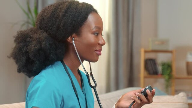 Close up shot of beautiful young African American woman nurse checking up measuring blood pressure of patient using tonometer and talking in good mood. Healthcare worker, social assistance, side view