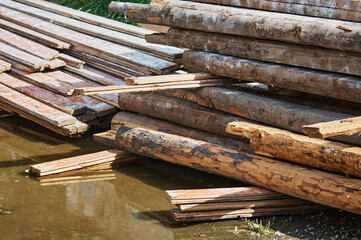 Timber on a construction site in a puddle after rain. Old wood of a demolition area. Hewn logs and old painted boards with tenon joints. Summer sunny day.