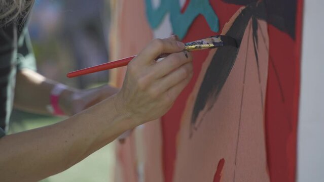 A handheld footage of a blonde woman's arms while painting on a white canvas while finishing the petals of a flower. She's holding her paint with her left hand while the other one holds the paintbrush