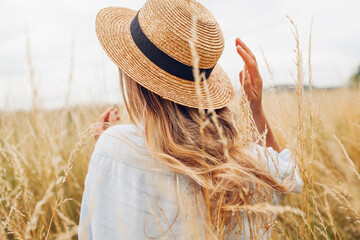 Back view of young woman walking among high grasses in summer meadow wearing straw hat and linen...