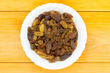 Raisins in a bowl. Dark dried grape in a deep white plate on an orange wooden table. Golden raisins. Brown and light raisins in a bowl. Traditional oriental sweets. Close-up. Copy space. Top view