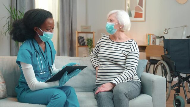 Portrait of Caucasian old lady in medical mask sitting on sofa in cozy living room listening to young pretty African American female nurse at home visit. Healthcare, medical consultation, quarantine