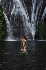 Fototapeta na wymiar Young athletic man swims in a mountain waterfall, Bali landscape, Indonesia. Tourism in Bali.
