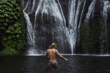 Naklejka na ściany i meble Young athletic man swims in a mountain waterfall, Bali landscape, Indonesia. Tourism in Bali.