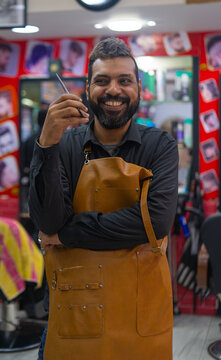 Attractive barber positive arab man smiling for camera and showing hairdressing scissors while working in vintage salon against barbershop background