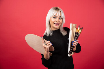 Young artist woman holding art supplies on a red background