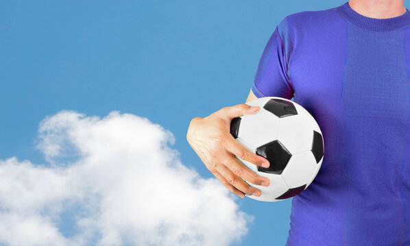 Cropped image of a young man holding a soccer ball with his hand wearing blue tshirt over isolated cloud background with copy space.Online cloud communication concept