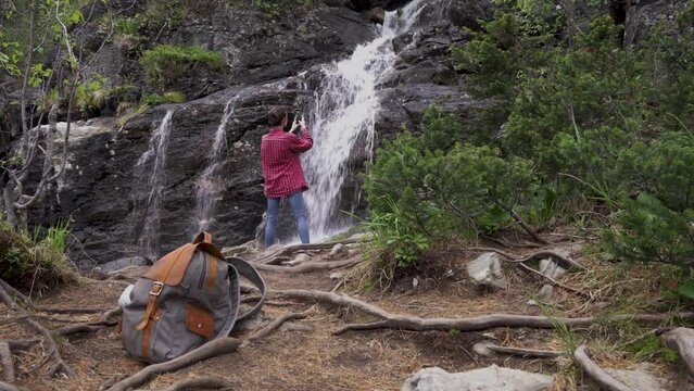 Young girl tourist with phone takes pictures of waterfall in forest. High quality 4k footage