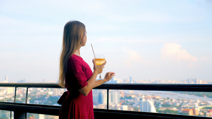 Young attractive woman drinking cocktail at luxury rooftop restaurant with high cityscape. Elegant female in red dress with mocktail at sky bar terrace enjoy rest with modern city skyline view.