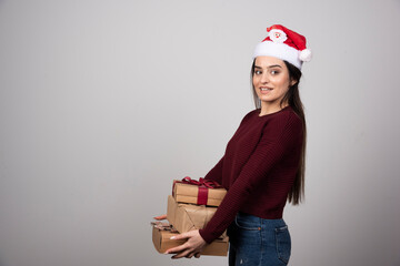 Young woman in Santa hat carrying Christmas presents on gray background