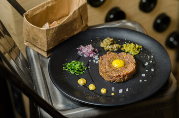 Gourmet tartar raw from beef fillet with yellow of the egg and vegetable as closeup. A dish on a metal chair against the background of bottles of wine. Wine bar menu. Restaurant