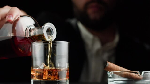 Drinking brandy or whiskey close-up. Rich man drinks luxury cognac in ice on black background. Alcohol amber rum, liqour beverage in glass. Smoking cigar. 