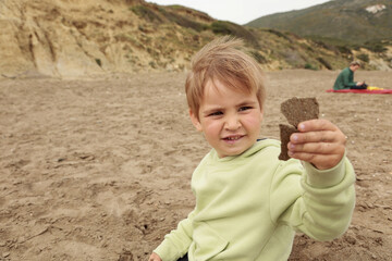 A little toddler 3years boy in yellow green hoodie is playing with sand on autumn spring beach cloudy day