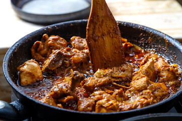 Chicken pieces stewed in sauce in spices in a deep frying pan with a wooden spatula.