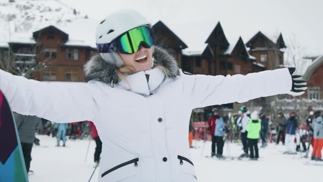 Happy young woman on ski slopes making a picture or video at ski resort in snowy mountains enjoying ski season vacations. Girl having fun in Aspen welcoming friends with wide opened hands, slow motion