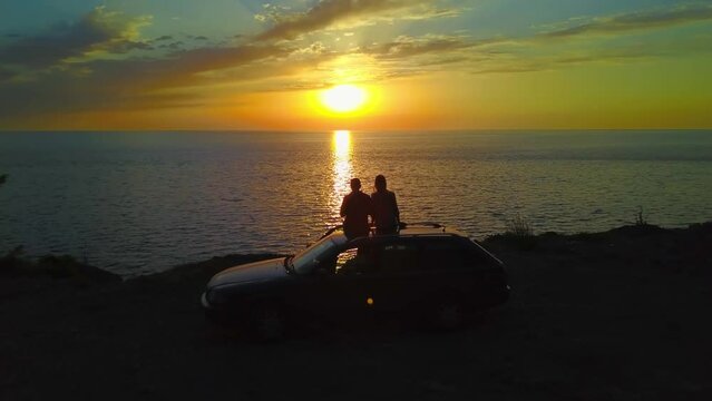 Shooting From Behind On Two People In Love, Couple Sits On Roof Of Car And Watch Sunset On Sea Or Ocean. Sky Is In Clouds, Drone And Camera Are In. Guy And Girl Escort Sun Over Horizon.