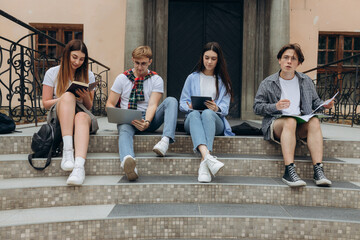 Fototapeta premium Study together is fun, teamwork, team building concept. Four happy students are sitting near college building and holding books, devices, wearing casual smart, smiling, on a nice summer