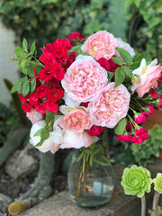 Bouquet of pink and red roses in a glass vase in a garden surrounded by flowers and plants