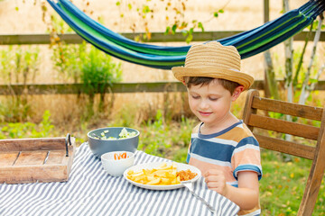 Little boy sit at table in a garden and eating  salad and french fries