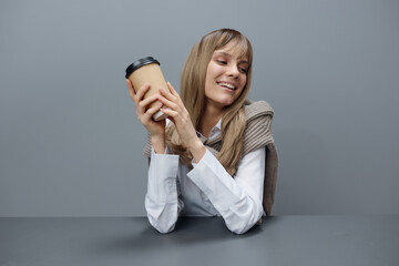 Cheerful cute young blonde student lady freelancer in warm sweater with takeaway coffee smiling looks aside in gray modern home office. Coffee break Lover Concept. Copy space. Cool offer