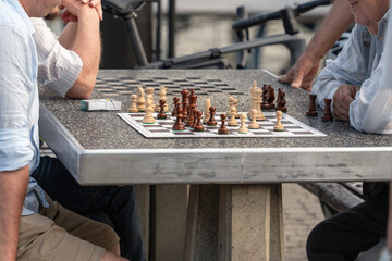 Chessboard with wooden chess pieces on a concrete street table, an elderly and a young man are...
