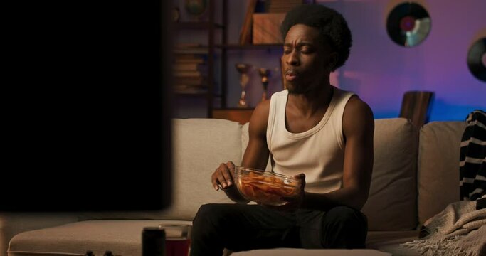 A Man With Dark Skin And An Afro Sits On The Couch In The Living Room Towering, The Guy Alone Watches His Favorite TV Series, Gets Excited About The Action, Eats A Snack Of Chips.