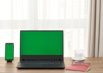 Fototapeta premium Laptop and mobile phone with green screen on wooden table against window background