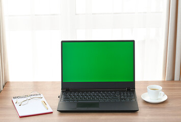 A computer with a green blank screen on a table with a notepad, glasses and a cup of coffee against the window.