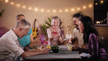 Group of merry friends laughing at an evening party toasting with cocktails and having fun together. Smiling people telling jokes enjoying the nightlife drinking alcoholic beverages.