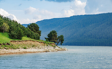 Deciduous trees grow on ledge above lake against backdrop of mountain range covered with vegetation...
