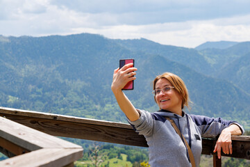Fototapeta na wymiar Woman stands on observation deck on high peak takes selfie on phone or shoots blog, against backdrop of mountain valley