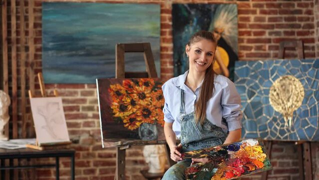 Portrait of young smiling female artist with her artworks in art studio. Painter holding art brushes and painting palette. Creative process, relaxation, leisure, hobby, stress management.