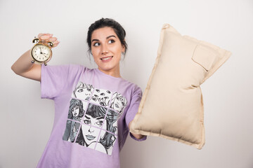 Young woman holding alarm clock and cushion on white background