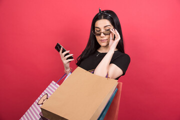 Young woman with glasses holding her bags and phone on red background