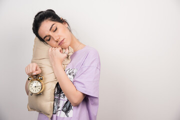 Sleepy woman holding alarm clock and cushion on white background