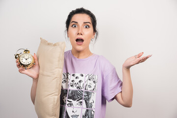 Shocked woman holding alarm clock and cushion on white background
