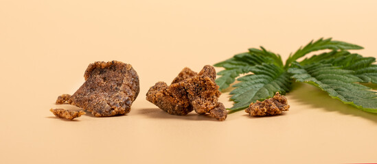 brown pieces of hashish and green cannabis leaf on a yellow background. - obrazy, fototapety, plakaty