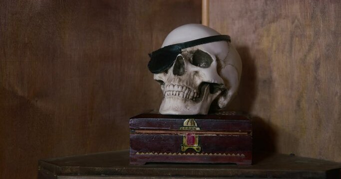 Fake or real human skull with a pirate eye patch stands on a wooden box on bedside table in the corner of the apartment, front view. Unusual thematic interior details