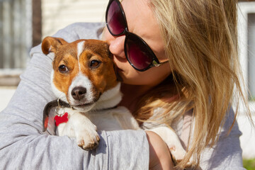 A young blonde woman in sunglasses hugs and kisses a funny dog jack russell terrier on a hot summer sunny day. Portrait, close-up. Love and care for animals concept