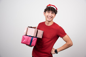 Young male in Santa hat holding gifts on gray background