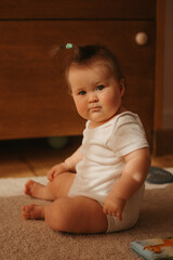 A 7-month girl is sitting near a dresser in a bodysuit at home. A serious infant in the sunlight.