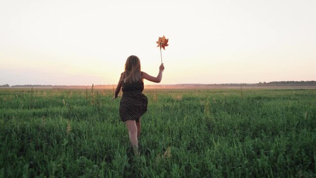 View from behind a child girl in a brown dress, running across a field with a toy windmill in her right hand. Concept of childhood, happiness and freedom. Peaceful life. Steadicam shot