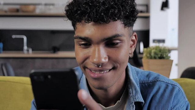 Young african american male holding smartphone device texting sms message sitting at home on sofa, smiling black guy using apps playing mobile game chatting in social media surfing web on phone
