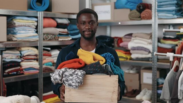 Portrait view of the multiracial male worker looking at the camera and stretching box with clothes while working at the donating center or second hand. Humanitarian aid concept