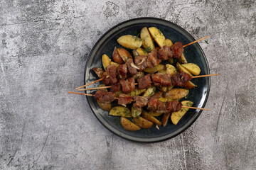 Kebabs on wooden skewers with potatoes and greens on a round plate on a dark gray background. Top view, flat lay