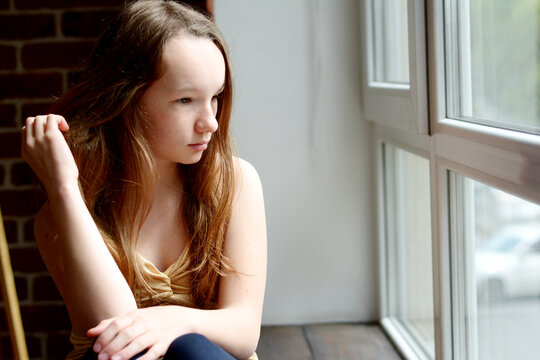 a teenage girl looks out the window she has long dark brown hair she is dressed in a golden t-shirt she thinks sadly about something with one hand she touches her hair. High quality photo