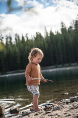 Little toddler girl with stick happily playing outdoors at wilderness lake