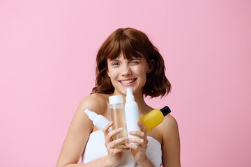 a young, well-groomed woman stands on a pink background holding different jars of care cosmetics in her hands and smiling broadly with her teeth, squints her eyes. Horizontal photo with an empty space