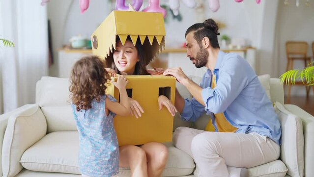 Happy family of parents and daughter child girl playing dinosaurs hunt at home, dad wearing handmade costume monster of cardboard chasing mom and daughter with fun and joyful exited in living room