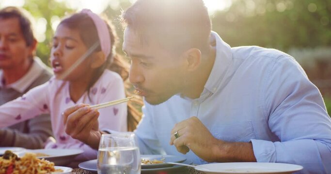 Young man using chopsticks to eat chinese food together with his family at a table outside. Having lunch or dinner with his father, daughter and other relatives at an event or gathering outdoors